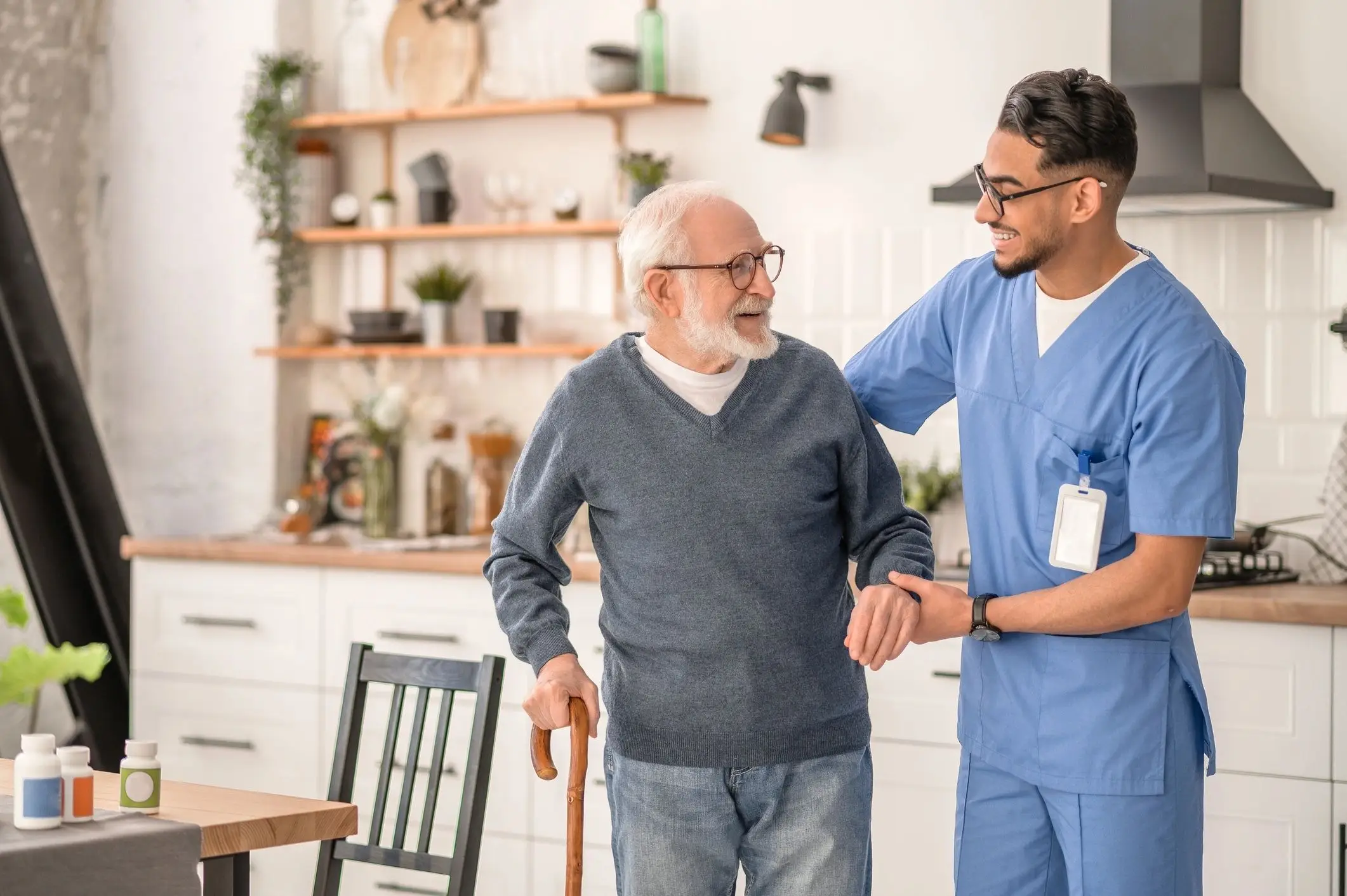 A caregiver in blue scrubs assists an elderly man with a cane in a bright, modern kitchen filled with plants and decor.