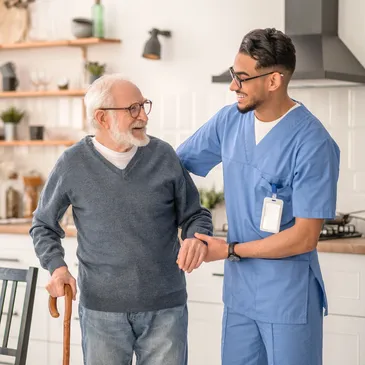 A caregiver assists an elderly man with a cane in a well-lit kitchen, featuring plants and shelves in the background.